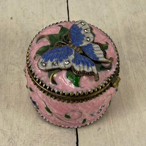 May include: A pink, oval-shaped trinket box with a detailed butterfly on the lid. The butterfly has blue wings with white accents and is adorned with small, clear gems. The box features a floral design and a bronze-colored trim.
