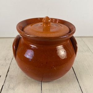 May include: A glazed, brown ceramic pot with a lid. The pot has a rounded body, two side handles, and a lid with a small knob. The ceramic has a glossy finish and a warm, earthy tone, suggesting it is vintage or handmade.