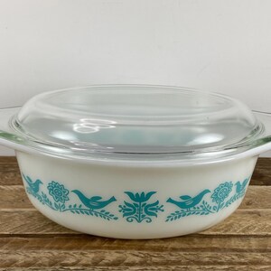 May include: White Pyrex casserole dish with a clear glass lid. The dish features a teal bird and floral pattern around the exterior. The dish has two handles and is sitting on a wooden surface.