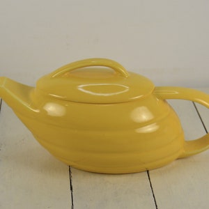 May include: A yellow ceramic teapot with a rounded body and a curved spout. The teapot has a lid with a handle and a separate handle on the side.