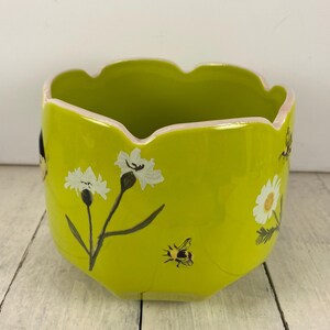 Może przedstawiać: Chartreuse ceramiczna donica z falistym brzegiem i kwiatowymi i pszczelimi wzorami. Donica posiada białe kwiaty z czarnymi łodygami i zielonymi liśćmi oraz małą pszczołę. Obrzeże jest pomalowane na r&oacute;żowo.