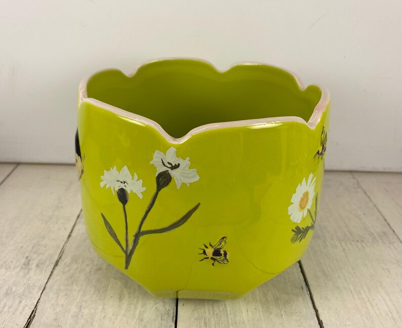 Może przedstawiać: Chartreuse ceramiczna donica z falistym brzegiem i kwiatowymi i pszczelimi wzorami. Donica posiada białe kwiaty z czarnymi łodygami i zielonymi liśćmi oraz małą pszczołę. Obrzeże jest pomalowane na r&oacute;żowo.