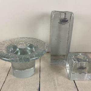 May include: A collection of clear glass candle holders. The set includes a round, shallow holder, a tall rectangular holder, and a square holder. Each piece has a textured surface and is designed to hold a single candle.