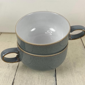 May include: Two stacked, gray ceramic soup bowls with handles. The bowls have a speckled texture and a light brown rim. The interior of the bowls is white. The bowls are on a white wooden surface.