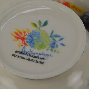 May include: A white ceramic bowl with a floral design in shades of blue, yellow, and orange. The bowl has the text "Anthropologie" and "Dishwasher & Microwave Safe" in black text. Made in China / Fabriqu&eacute; en Chine.