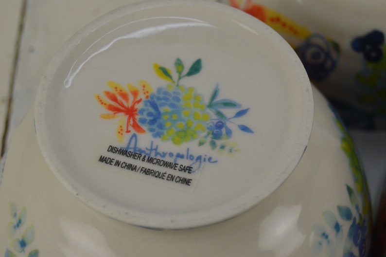 May include: A white ceramic bowl with a floral design in shades of blue, yellow, and orange. The bowl has the text "Anthropologie" and "Dishwasher & Microwave Safe" in black text. Made in China / Fabriqu&eacute; en Chine.