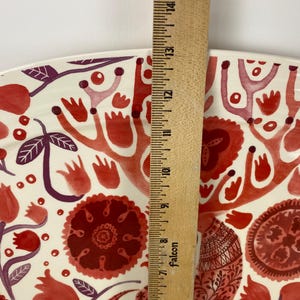 Op de afbeelding: Een decoratieve keramische schaal met een bloemmotief in tinten rood en paars op een cr&egrave;mekleurige achtergrond. De schaal wordt gemeten met een houten liniaal, met een lengte van ongeveer 35,5 cm.