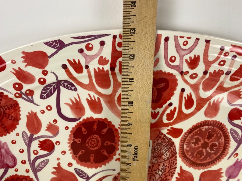Op de afbeelding: Een decoratieve keramische schaal met een bloemmotief in tinten rood en paars op een cr&egrave;mekleurige achtergrond. De schaal wordt gemeten met een houten liniaal, met een lengte van ongeveer 35,5 cm.