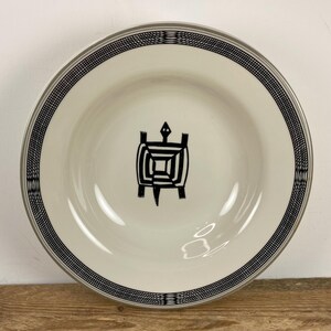 May include: A round, off-white ceramic bowl featuring a centered black line drawing of a turtle. The wide rim is decorated with a detailed black cross-hatched pattern that encircles the entire edge of the bowl, creating a distinct contrast.