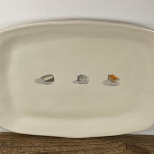 May include: Cream-colored rectangular serving platter with rounded edges. The platter features three small illustrations of food items: a wedge of cheese labeled "brie", a wheel of cheese, and a rectangular block of cheese labeled "cheddar".