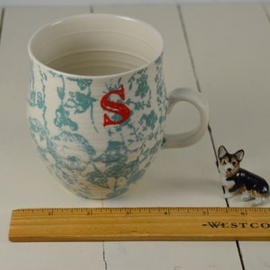 May include: A white ceramic mug with a blue and white floral pattern and the letter "S" in red. The mug is sitting on a white wooden surface.