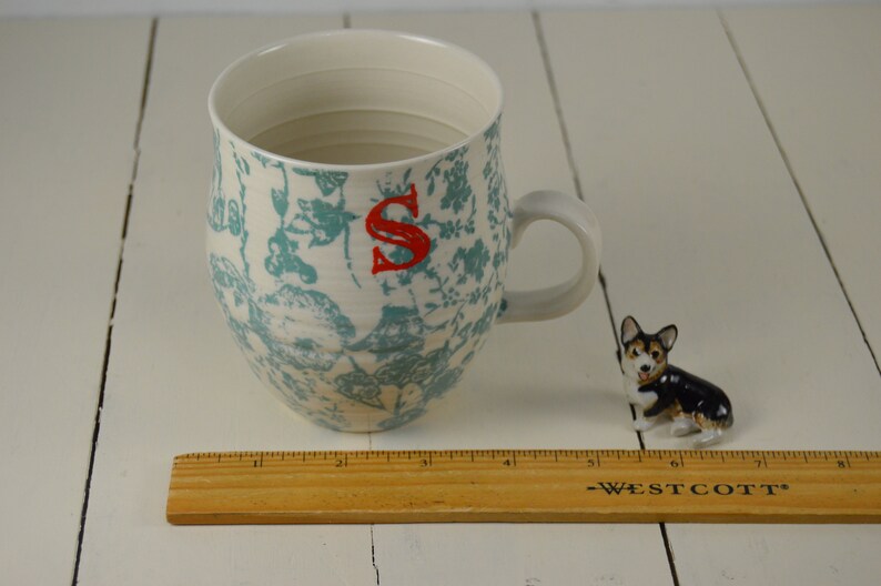 May include: A white ceramic mug with a blue and white floral pattern and the letter "S" in red. The mug is sitting on a white wooden surface.