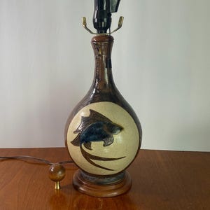 May include: A ceramic table lamp with a dark brown, mottled glaze and a central cream-colored circular medallion featuring a dark blue and brown incised fish design. The lamp has a wooden base, a black socket, and a separate small wooden knob.