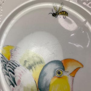 May include: White ceramic plate with a painted bird and a bee. The bird has yellow, blue, and orange details. The bee is yellow and black. The plate has a decorative rim.