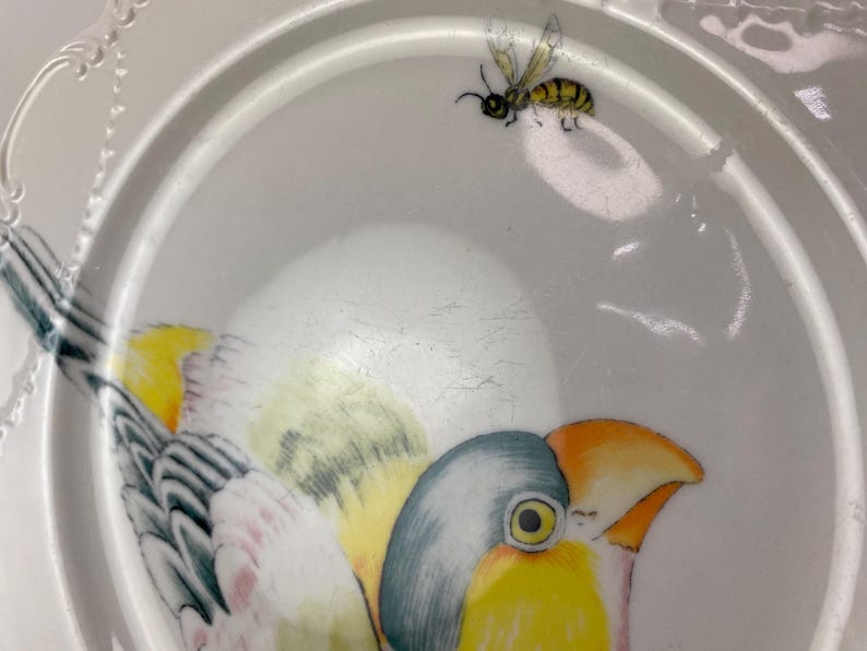 May include: White ceramic plate with a painted bird and a bee. The bird has yellow, blue, and orange details. The bee is yellow and black. The plate has a decorative rim.