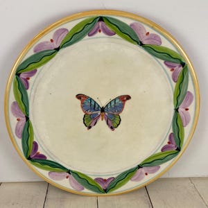 May include: Round ceramic plate with a cream-colored center featuring a colorful butterfly. The rim is decorated with a border of green leaves and purple flowers, with a gold-colored edge.