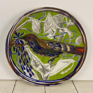 K&ouml;nnte beinhalten: Dekorative Keramikplatte mit einem leuchtend gr&uuml;nen Hintergrund und einer detaillierten Illustration eines Vogels auf einem Ast. Der Vogel hat braunes, rotes und blaues Gefieder. Die Platte hat einen goldenen Rand, einen blau-wei&szlig;en Rand und einen Durchmesser von ca. 25 cm.