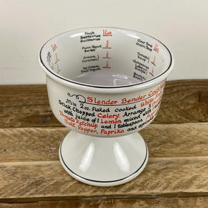 May include: White ceramic measuring cup with a pedestal base and black trim. The cup features red and black text indicating measurements and recipes, including "Slender Bender Cocktail". The cup is designed for kitchen use.