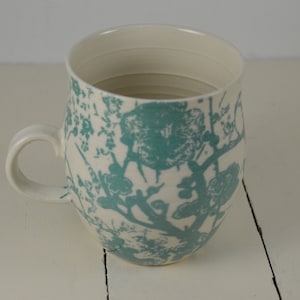 May include: A white ceramic mug with a teal floral pattern. The mug has a rounded body and a handle.