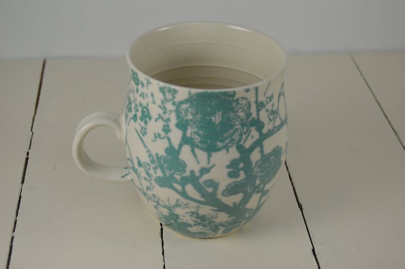 May include: A white ceramic mug with a teal floral pattern. The mug has a rounded body and a handle.