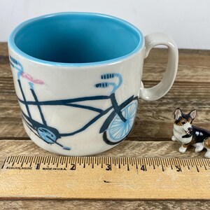 K&ouml;nnte beinhalten: Eine Keramiktasse mit blauem Innenraum und einem Fahrradmuster in Schwarz und Blau. Neben der Tasse befindet sich eine kleine Corgi-Figur. Ein Holzlineal mit Zollma&szlig;en befindet sich im Vordergrund.