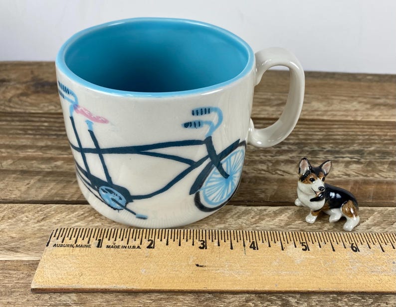 K&ouml;nnte beinhalten: Eine Keramiktasse mit blauem Innenraum und einem Fahrradmuster in Schwarz und Blau. Neben der Tasse befindet sich eine kleine Corgi-Figur. Ein Holzlineal mit Zollma&szlig;en befindet sich im Vordergrund.