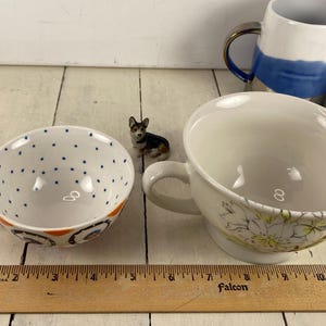 K&ouml;nnte beinhalten: Zwei Keramik-Teetassen mit Henkeln, eine wei&szlig; mit blauen Punkten und orangefarbenen Akzenten, die andere wei&szlig; mit floralem Design. Zwischen ihnen sitzt eine kleine Corgi-Figur. Ein Holzlineal befindet sich im Vordergrund.