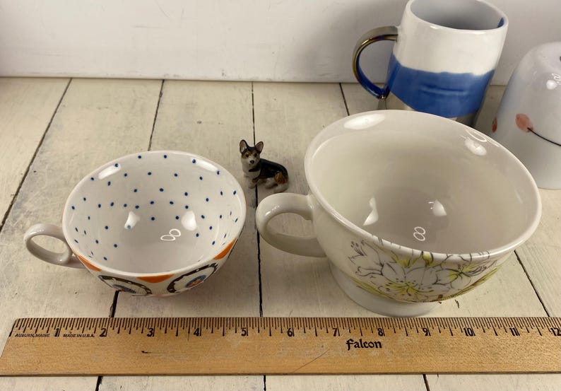 K&ouml;nnte beinhalten: Zwei Keramik-Teetassen mit Henkeln, eine wei&szlig; mit blauen Punkten und orangefarbenen Akzenten, die andere wei&szlig; mit floralem Design. Zwischen ihnen sitzt eine kleine Corgi-Figur. Ein Holzlineal befindet sich im Vordergrund.