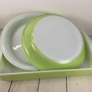 May include: A set of three vintage Pyrex baking dishes in a light green color. The set includes a rectangular dish and two round dishes, one of which is inverted. The dishes have white interiors and are stacked together.