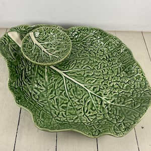 May include: A glossy green ceramic serving platter shaped like a large cabbage leaf with a smaller, integrated circular bowl attached to the top left side. The surface features a highly detailed, embossed texture mimicking natural leaf veins.