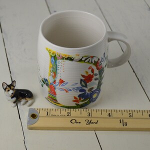 May include: White ceramic mug with a floral design and a small corgi figurine. The mug has a curved handle and a colorful floral pattern. A wooden ruler is next to the mug, measuring in inches.