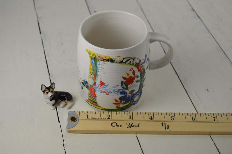 May include: White ceramic mug with a floral design and a small corgi figurine. The mug has a curved handle and a colorful floral pattern. A wooden ruler is next to the mug, measuring in inches.