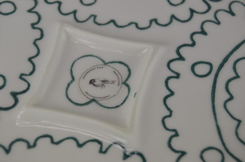 K&ouml;nnte beinhalten: Wei&szlig;e Keramikplatte mit gr&uuml;nem Dekorrand. Die Platte hat einen kleinen, separaten Abschnitt mit einem gr&uuml;nen Kleeblatt-Design und dem Text "Johansson Sala White Edition" im Inneren.