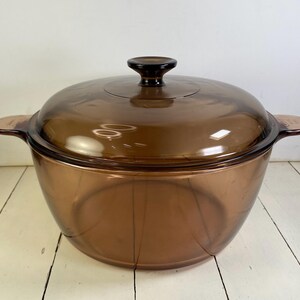 May include: A round, transparent amber-colored glass cooking pot with a matching lid. The pot features two integrated side handles and a central knob on the lid. The glass is smooth and uniform in color, allowing for a clear view through the vessel.