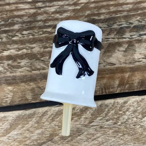 May include: A white popsicle mold with a black bow design. The popsicle mold has a light-colored stick and is set against a wooden background.