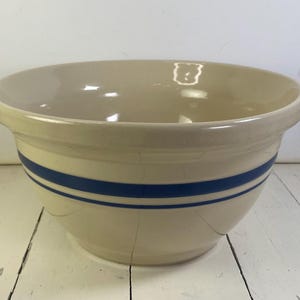 May include: A large, round, cream-colored ceramic mixing bowl featuring a wide, dark blue horizontal stripe encircling the exterior. The bowl has a smooth, glossy finish, a thick rim, and a slightly tapered base, showing visible surface cracks.