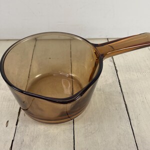 May include: A vintage amber-colored glass saucepan with a handle. The saucepan has a rounded shape and a pouring spout. The handle is rectangular and extends from the side of the pan. The glass is transparent, allowing the contents to be seen.