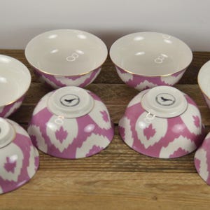 May include: Set of six white ceramic bowls with pink and white ikat pattern. The bowls have a gold rim and a small bird logo on the bottom.