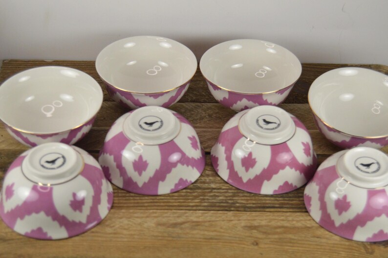 May include: Set of six white ceramic bowls with pink and white ikat pattern. The bowls have a gold rim and a small bird logo on the bottom.