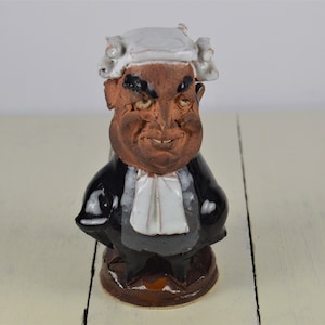 May include: A ceramic figurine of a judge wearing a black robe and a white wig. The figurine has a brown face and a serious expression.