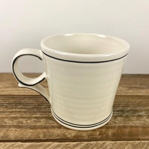 Puede incluir: Taza de cer&aacute;mica color crema con un borde negro alrededor del borde y la base, y un contorno negro en el asa. La taza est&aacute; sobre un fondo claro y descansa sobre una superficie de madera.