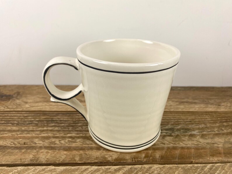 Puede incluir: Taza de cer&aacute;mica color crema con un borde negro alrededor del borde y la base, y un contorno negro en el asa. La taza est&aacute; sobre un fondo claro y descansa sobre una superficie de madera.