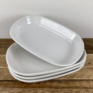May include: A stack of four white ceramic oval dishes with smooth, glossy surfaces and slightly raised edges. The dishes are uniform in shape and color, featuring a simple, clean design with a shallow basin and rounded corners for serving food.