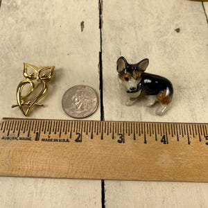 Op de afbeelding: Een goudkleurige uilvormige broche en een klein keramisch hondenbeeldje met zwarte, bruine en witte markeringen worden getoond. Een munt en een houten liniaal zijn ook aanwezig, wat de grootte van de items aangeeft.