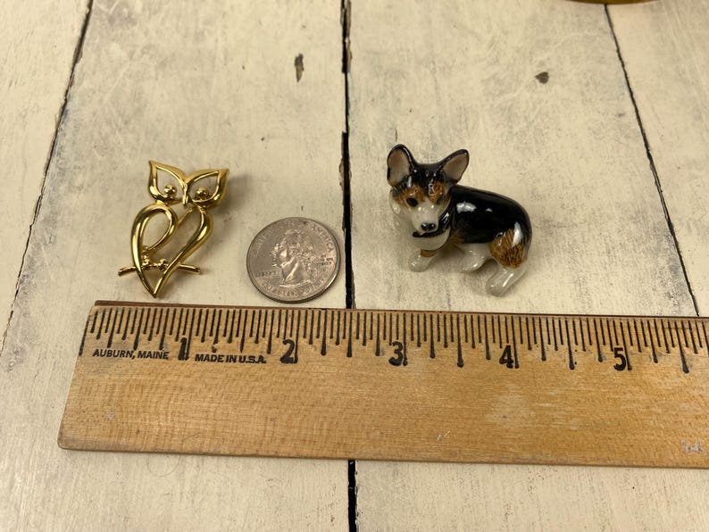 Op de afbeelding: Een goudkleurige uilvormige broche en een klein keramisch hondenbeeldje met zwarte, bruine en witte markeringen worden getoond. Een munt en een houten liniaal zijn ook aanwezig, wat de grootte van de items aangeeft.