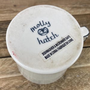 Puede incluir: Taza de cer&aacute;mica blanca con exterior texturizado y asa. La base de la taza presenta el texto "molly hatch" en escritura azul, con un dise&ntilde;o de corona. Una etiqueta indica que la taza es apta para lavavajillas y microondas.
