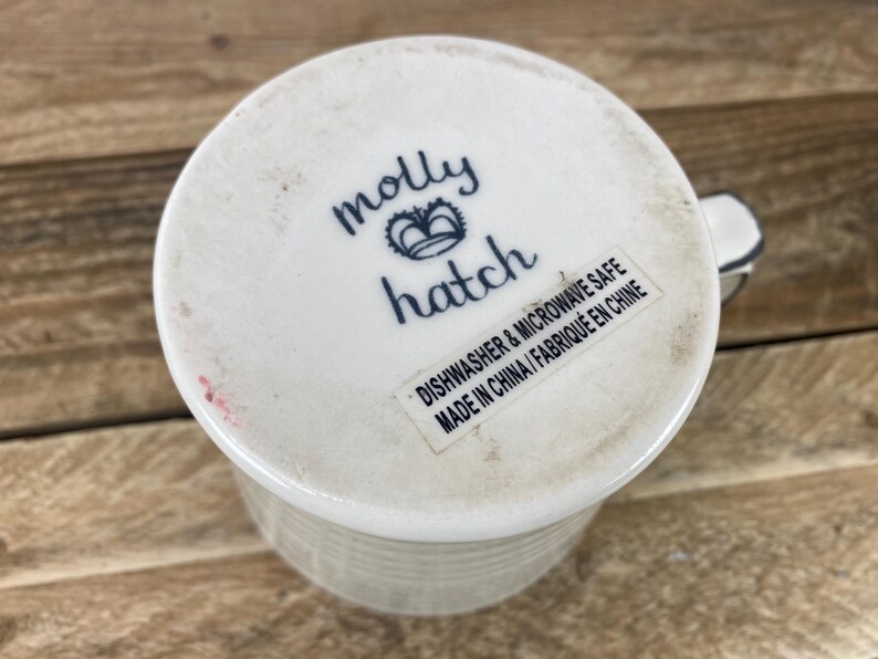 Puede incluir: Taza de cer&aacute;mica blanca con exterior texturizado y asa. La base de la taza presenta el texto "molly hatch" en escritura azul, con un dise&ntilde;o de corona. Una etiqueta indica que la taza es apta para lavavajillas y microondas.