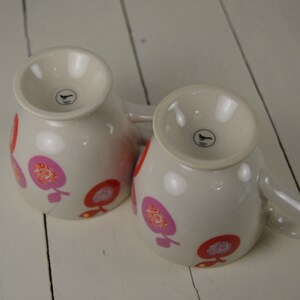 May include: Two white ceramic mugs with pink and red floral designs. The mugs have a black bird logo on the bottom.