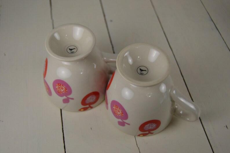 May include: Two white ceramic mugs with pink and red floral designs. The mugs have a black bird logo on the bottom.