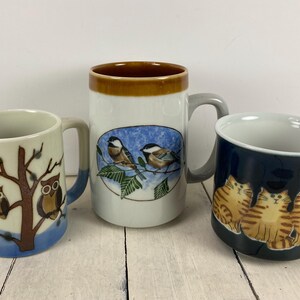 May include: Three ceramic mugs with animal designs. One mug features owls on a tree branch, another has birds on a branch, and the third has cats. Each mug has a handle and a unique color scheme.